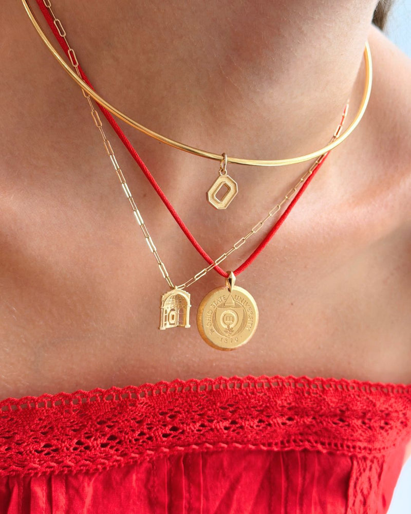 A close-up of a person wearing layered necklaces: a gold chain choker with a small geometric pendant, a gold chain with a small bird pendant, and a red cord necklace with a round gold medallion pendant.