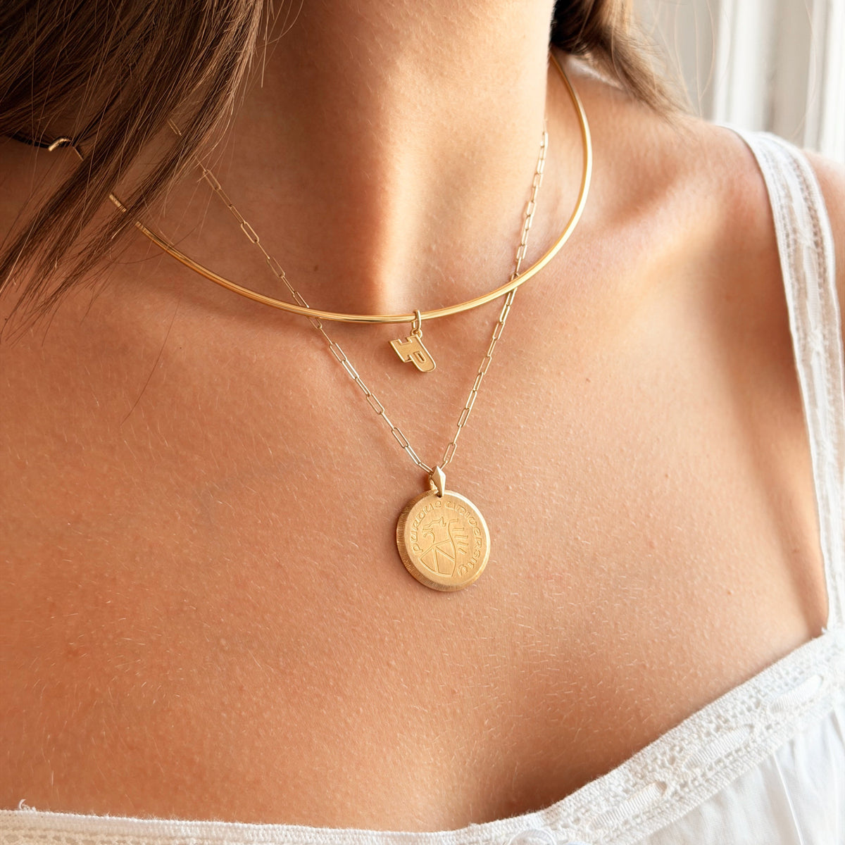 A close-up of a model wearing a layered gold necklace set, featuring a thin curved chain with a small lightning-bolt charm and a longer chain with a round coin pendant.