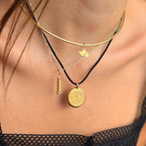 A close-up of a model wearing a layered necklace set. The set includes a gold-tone double-helix choker, a gold-tone lightning bolt charm, a gold-tone cylinder charm, and a gold-tone coin pendant on a black cord, layered over a thin silver-tone chain.