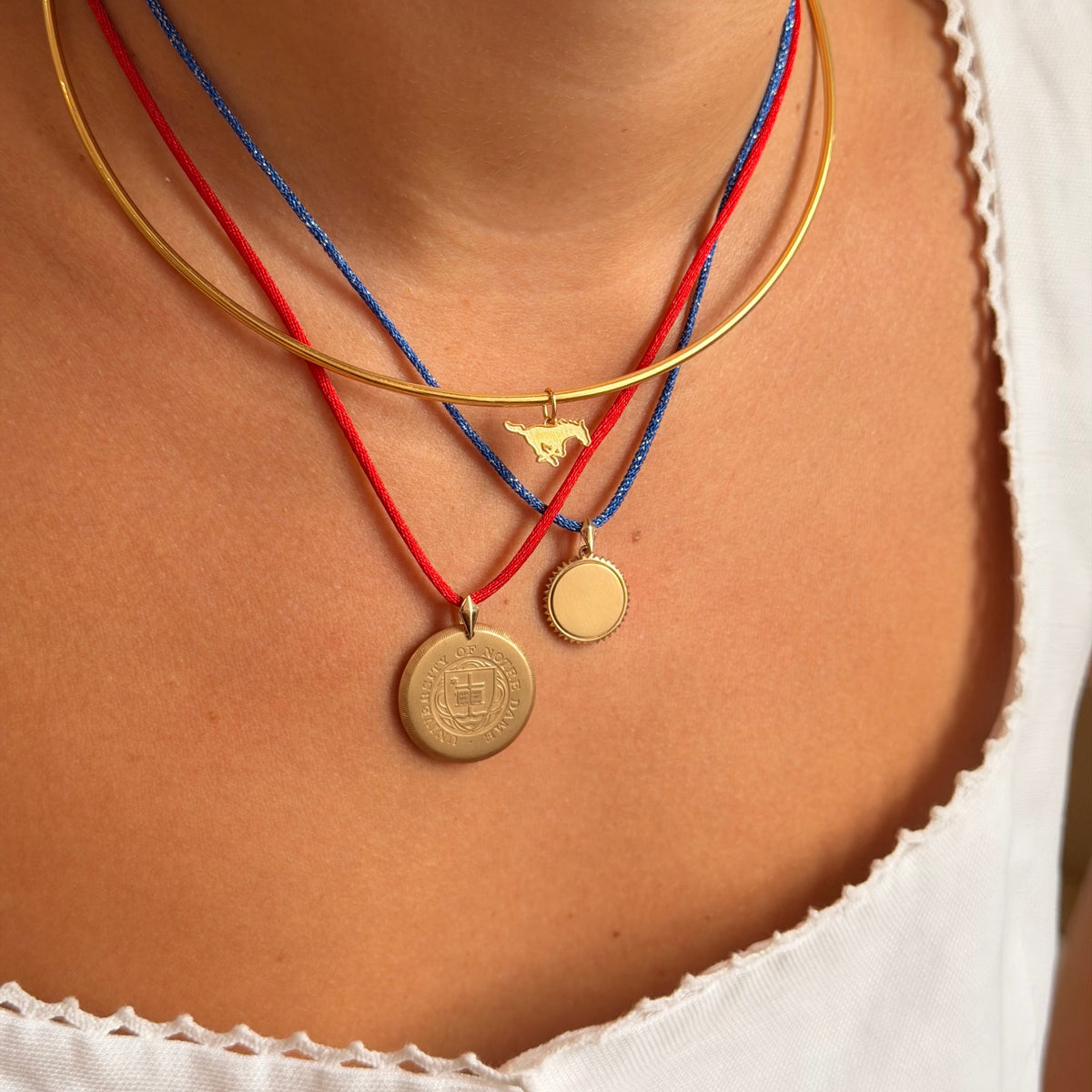 A close-up of a person's neck and chest wearing a layered necklace set. The set includes a gold bangle, a red cord necklace with a small gold bird charm, a blue cord necklace with a small round gold medallion, and a gold coin-style medallion on a red cord.
