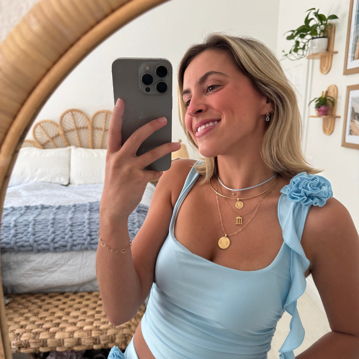 A woman takes a mirror selfie in a bedroom while wearing a light blue ribbed crop top and matching bottoms. The top features ruffled shoulder details and side ties, and she is wearing layered gold necklaces.