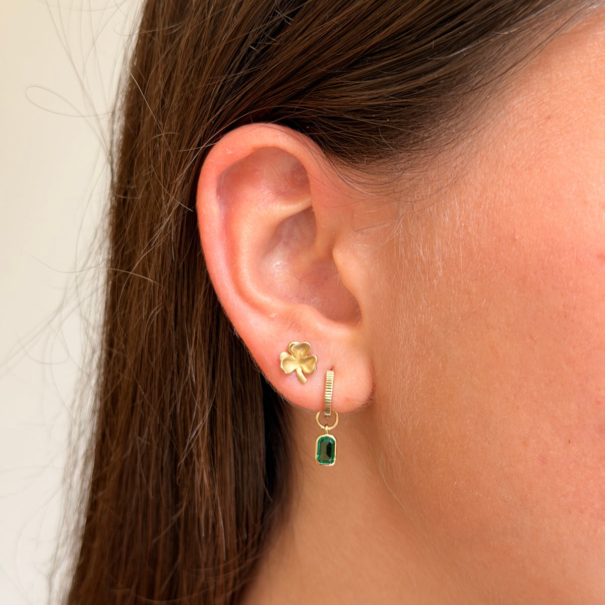 A close-up of a person's ear wearing a gold hoop earring with a small gold four-leaf clover stud and a dangling gold charm with a green rectangular stone.