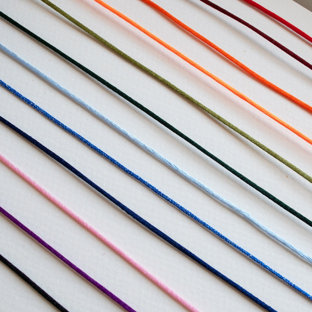 A close-up of multiple thin cords or threads arranged diagonally on a white background, shown in several colors including black, purple, pink, blue, green, orange, and red.