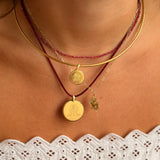 A close-up of a person's neck and upper chest wearing layered necklaces: a gold chain, a thin gold bangle, a red beaded necklace, and a red cord necklace with round gold pendants and a small gold leaf charm.