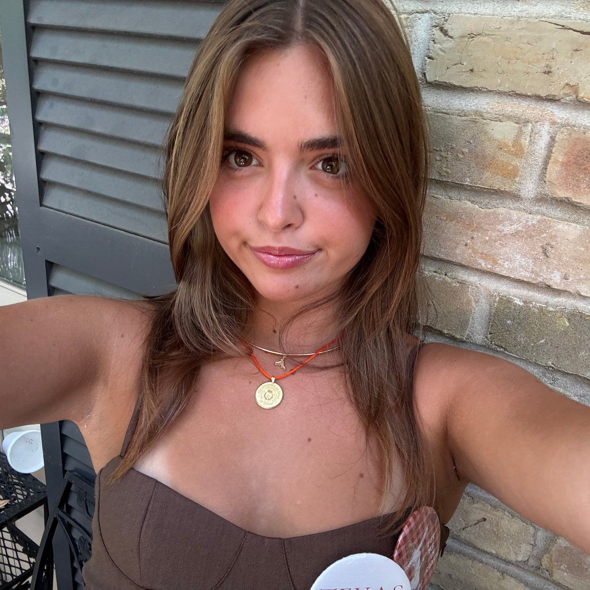 A woman taking a selfie outdoors, wearing a brown strapless top and layered necklaces, standing in front of a brick wall.