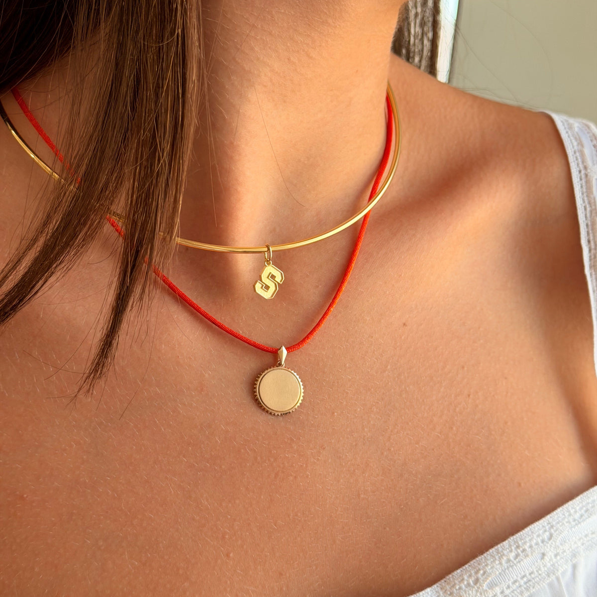 A close-up of a model wearing a double-strand necklace with a gold bangle-style top layer and a red cord bottom layer, both featuring small gold pendants.
