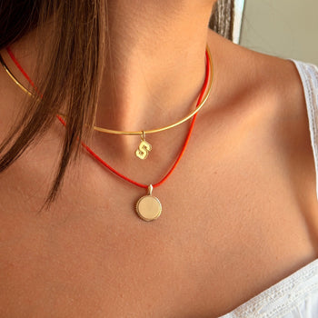 A close-up of a model wearing a double-strand necklace with a gold bangle-style top layer and a red cord bottom layer, both featuring small gold pendants.