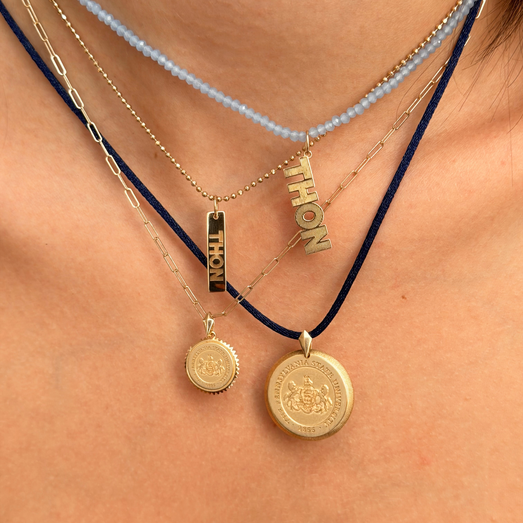 Layered necklaces worn on a person's neck, featuring gold-tone pendants including a baroque chain, a beaded light blue chain, a ball chain with a vertical 'THION' pendant, and two round gold medallion pendants on a dark cord.
