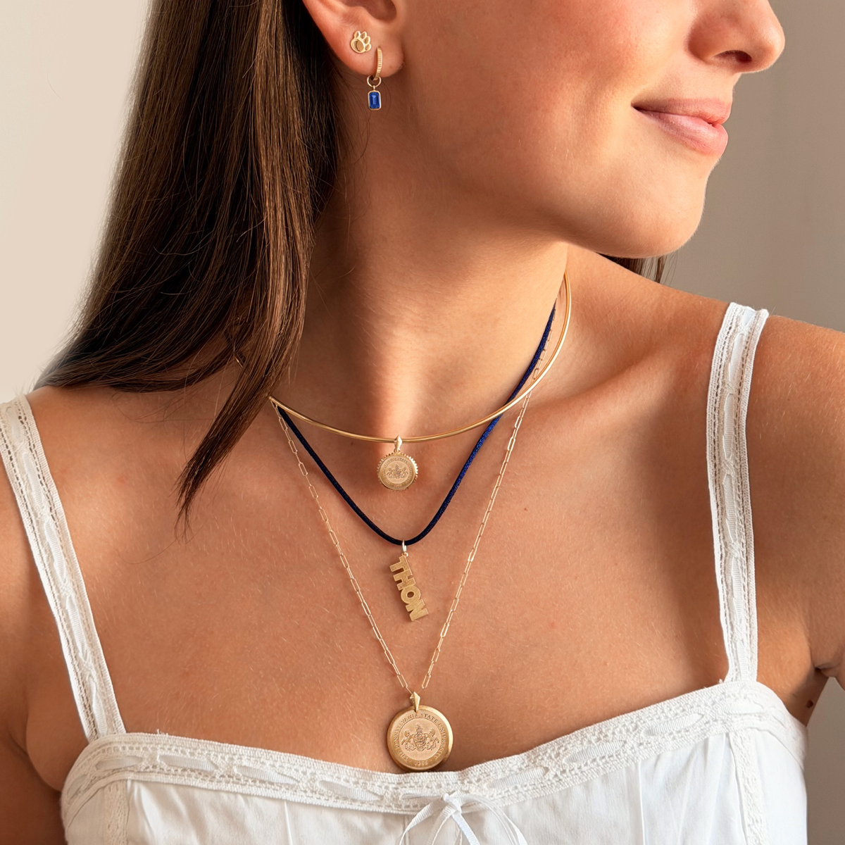 A close-up of a woman wearing layered necklaces, including gold coin pendants, a gold nameplate pendant spelling 'LOVE', and a dark blue cord necklace, along with a small gold and blue earring.