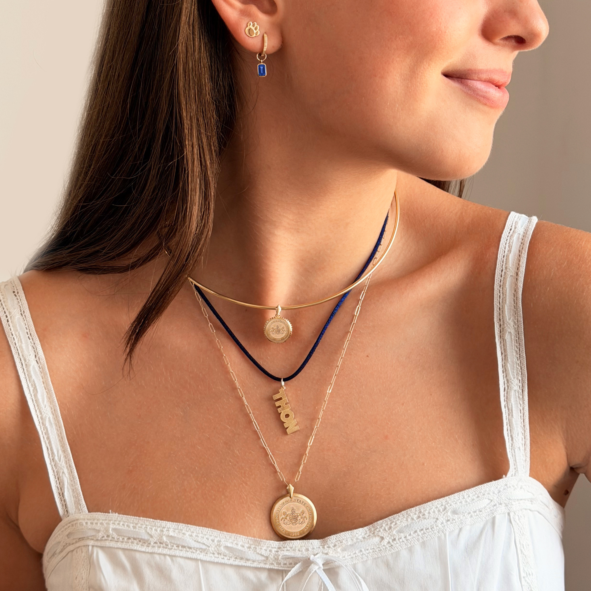 A close-up of a woman wearing layered necklaces, including gold coin pendants, a gold nameplate pendant spelling 'LOVE', and a dark blue cord necklace, along with a small gold and blue earring.