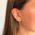 A close-up of a person's ear wearing a small gold hoop earring with a dangling rectangular amber-orange stone and a tiny gold letter T stud on the other ear.