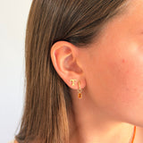 A close-up side profile of a woman wearing a delicate gold huggie hoop earring with a small rectangular amber gemstone dangle.