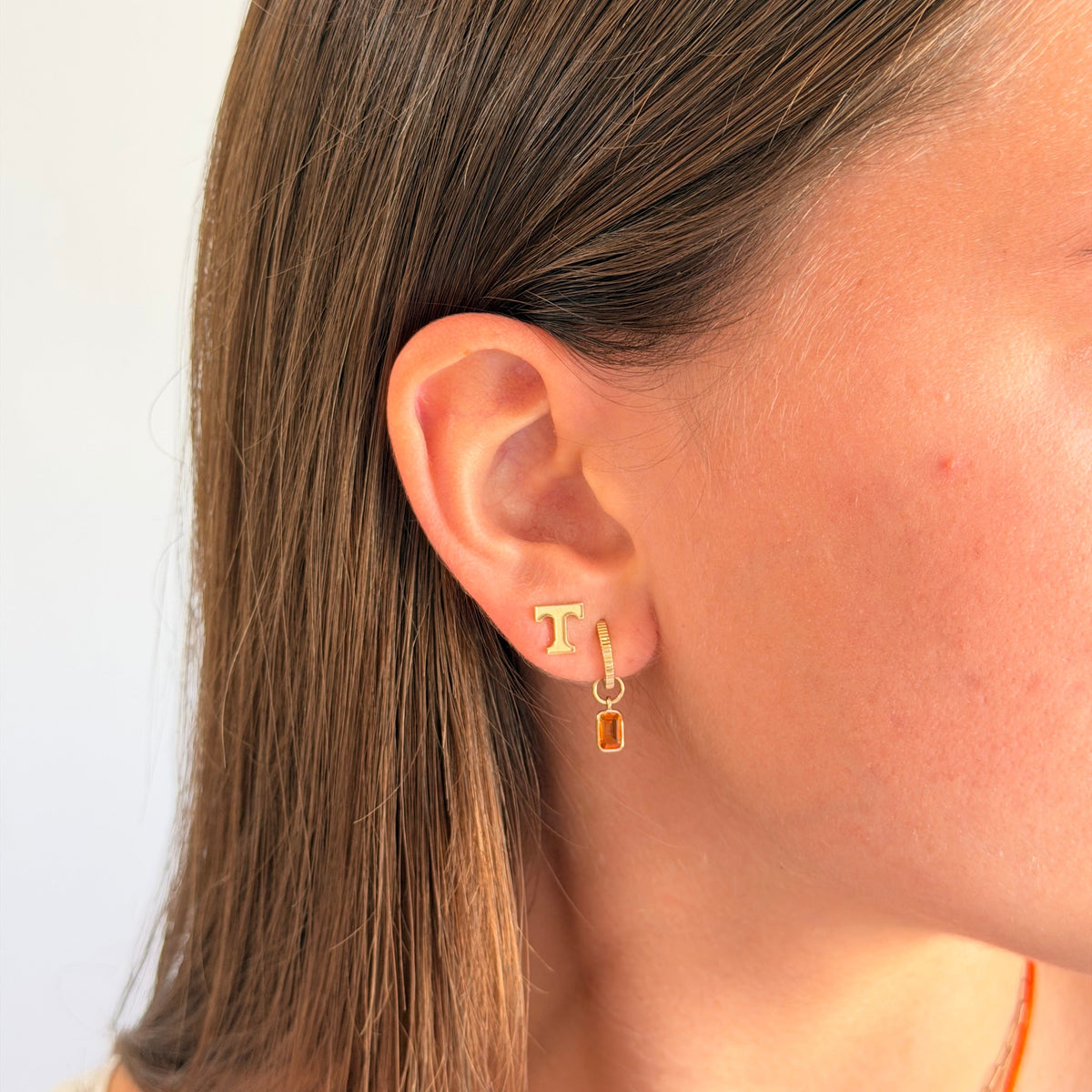 A close-up side profile of a woman wearing a delicate gold huggie hoop earring with a small rectangular amber gemstone dangle.