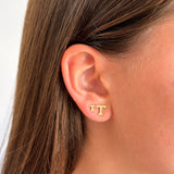 A close-up of a person's ear wearing a small gold stud earring with the letters 'TT' in a blocky font, against a light neutral background.