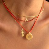 A close-up of a person's neck and chest wearing layered necklaces: a gold bangle, a red beaded chain with a small gold pendant, a gold chain with a dangling gold pendant, and a red cord with a gold pendant.