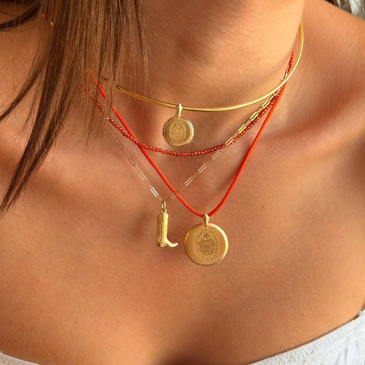 A close-up of a model's neck and chest wearing a layered necklace set. The set includes a gold bangle choker, a red cord necklace with a small gold boot charm, a red cord necklace with a round gold medallion, and a silver chain with a small gold charm.