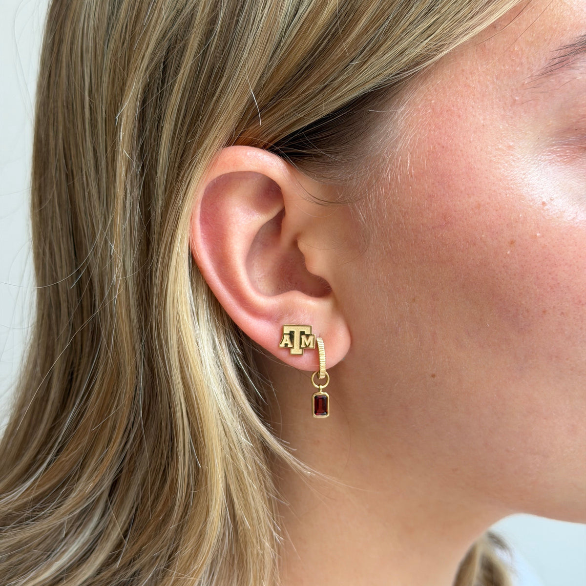 A close-up of a woman's ear wearing a gold stud earring with a small 'M' charm and a dangling rectangular dark red gemstone.
