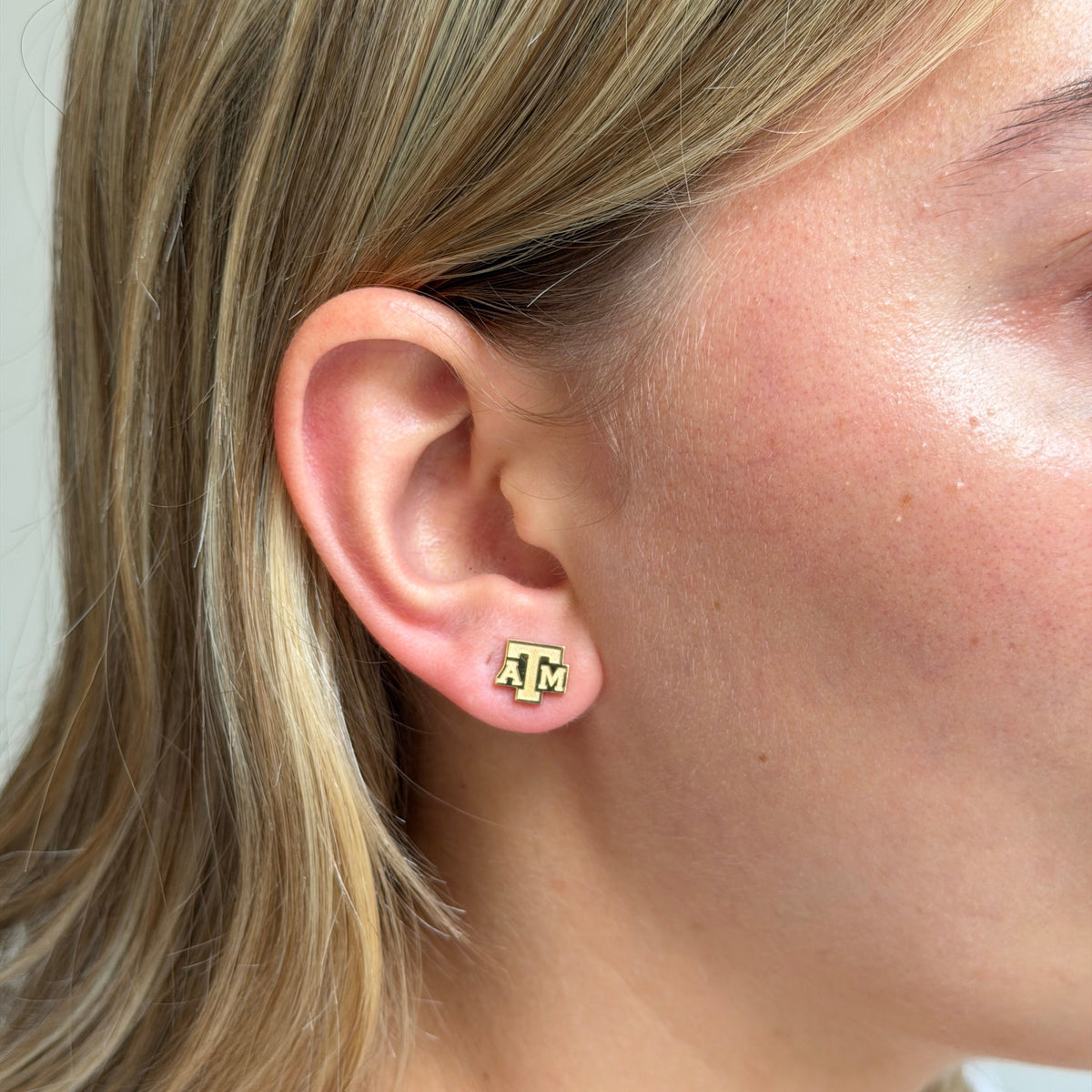 A close-up of a person's ear wearing a small gold stud earring with the Texas A&M 'A&M' logo.