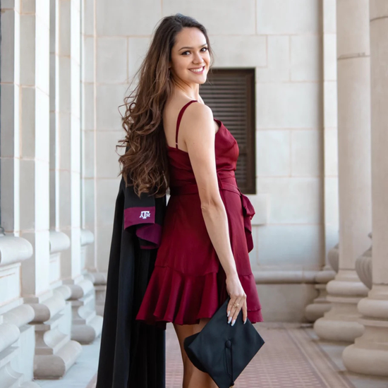 Woman in a red dress with a black cape holding a graduation cap, standing in an architectural setting.