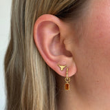 A close-up of a woman's ear wearing a gold hoop earring with a small gold Texas longhorn symbol and a dangling rectangular amber gemstone.