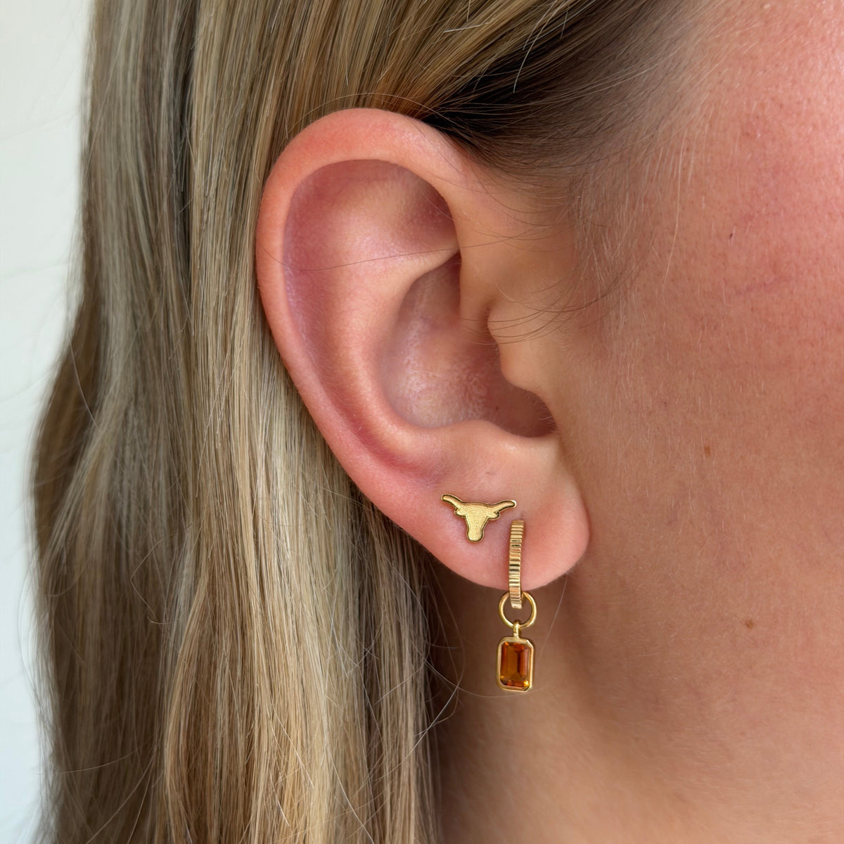 A close-up of a woman's ear wearing a gold hoop earring with a small gold Texas longhorn symbol and a dangling rectangular amber gemstone.