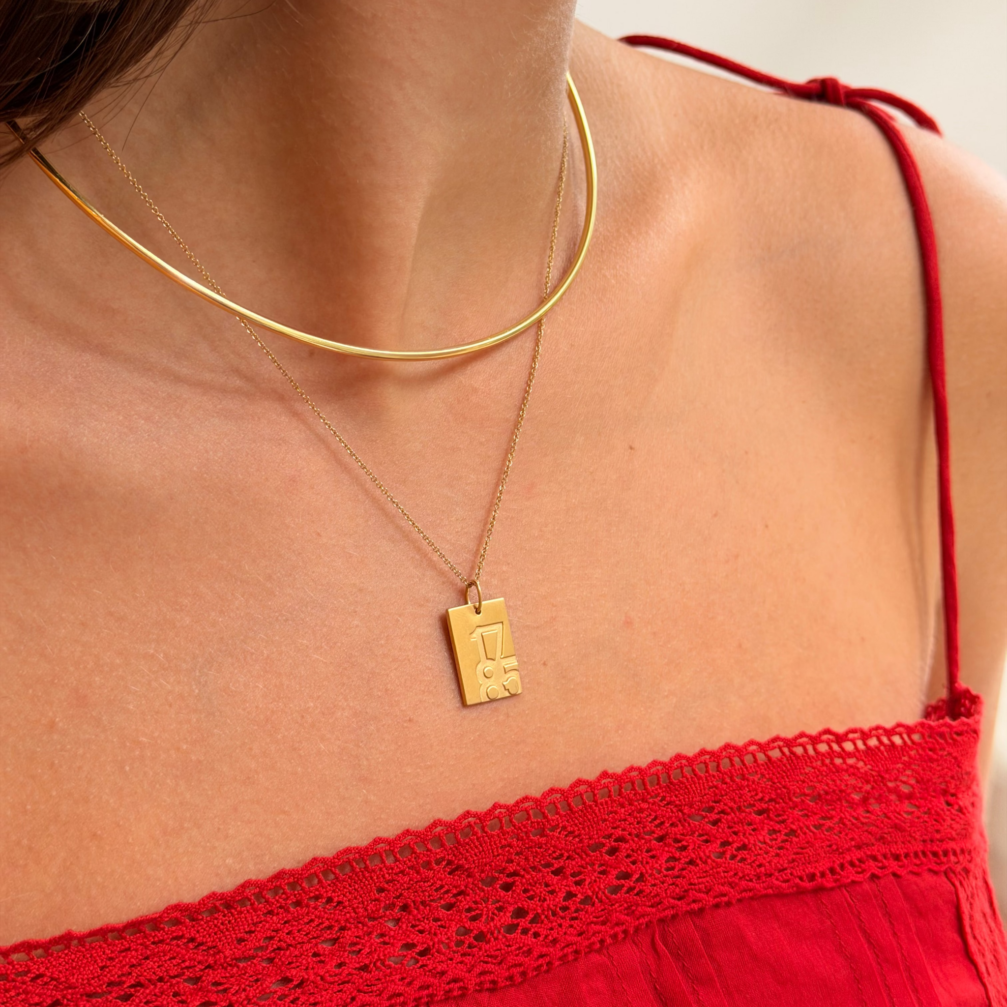 A close-up of a model wearing a gold necklace with a rectangular pendant featuring a stylized letter 'L', layered with a thin gold chain choker. The model is wearing a red lace-strap top against a light neutral background.