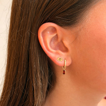 A close-up of a woman's ear wearing a small gold hoop earring with a dangling rectangular red gemstone, shown against her hair and skin.