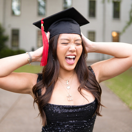 Graduate in cap and gown with red tassel celebrating outdoors.