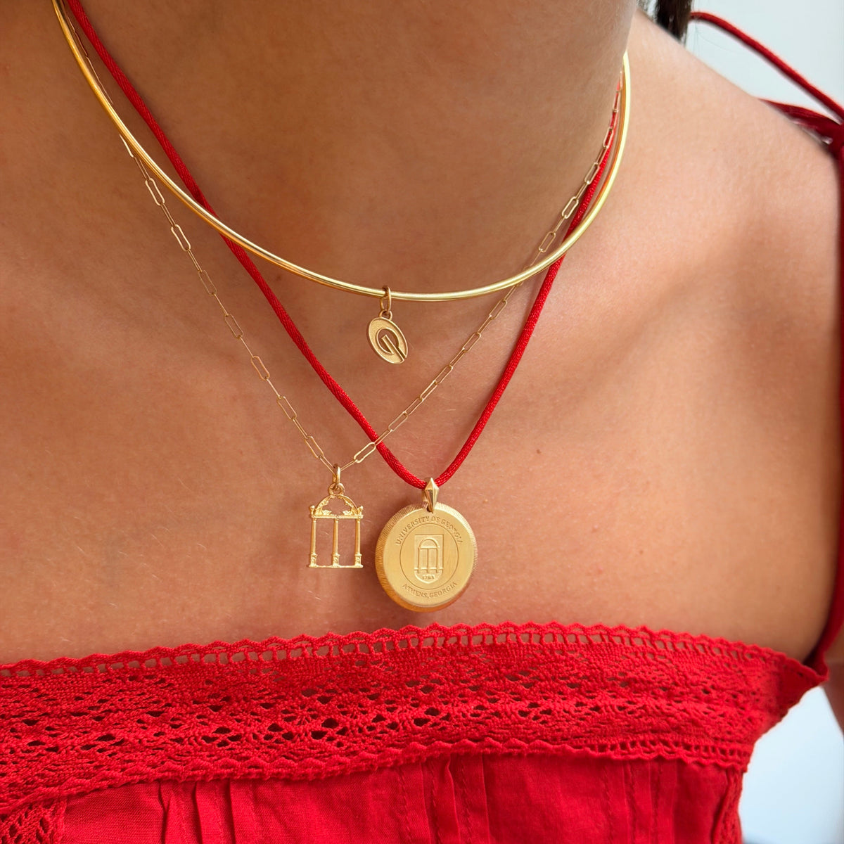 A close-up of a person's neck and chest wearing layered necklaces: a gold chain with a small crescent pendant, a gold chain with a small birdcage pendant, and a red cord necklace with a round gold pendant. The person is wearing a bright red off-the-shoulder top.