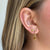 A close-up of a person's ear wearing a small gold huggie hoop earring with a dangling gold charm featuring a rectangular yellow gemstone.