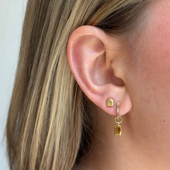 A close-up of a person's ear wearing a small gold huggie hoop earring with a dangling gold charm featuring a rectangular yellow gemstone.