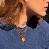 A close-up lifestyle shot of a woman wearing layered necklaces, featuring a prominent gold circular pendant on an orange cord, layered with a thin gold chain and a blue beaded necklace.