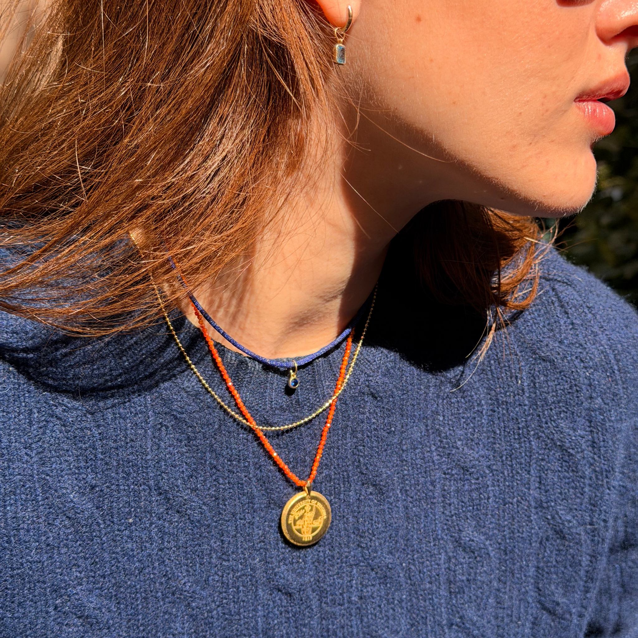 A close-up lifestyle shot of a woman wearing layered necklaces, featuring a prominent gold circular pendant on an orange cord, layered with a thin gold chain and a blue beaded necklace.