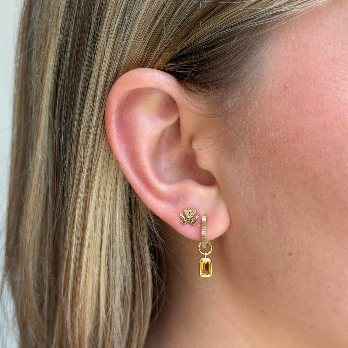 A close-up of a person's ear wearing a small gold huggie hoop earring with a dangling yellow rectangular charm and a matching gold leaf stud earring.