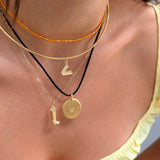 A close-up of a person's neck and upper chest wearing a layered necklace stack. The stack includes a thin gold chain, a textured orange beaded strand, a gold bangle, a black cord with a small gold geometric charm, a gold chain with a boot-shaped pendant, and a round gold coin pendant.