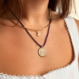A close-up of a person's neck and chest wearing layered necklaces: a gold bangle necklace with a small bird charm, and a dark blue cord necklace with a round gold pendant.