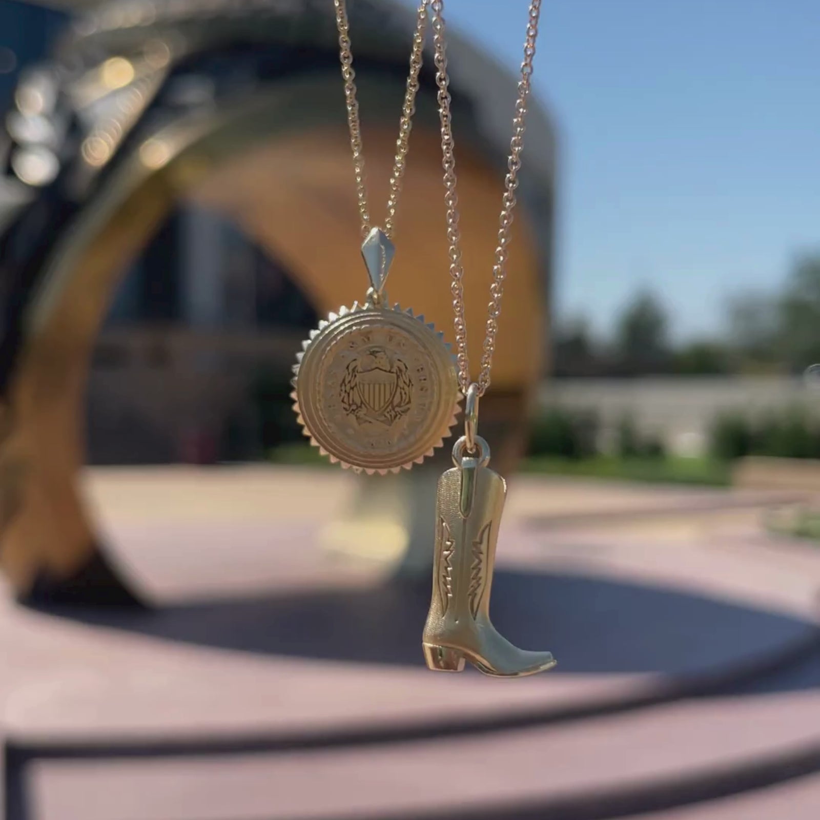 Texas A&M Former Students Sunburst Pendant shown in gold in size Standard on Cable Chain with Cowboy Boot Charm in gold on Cable Chain in front of Texas A&M Ring Statue